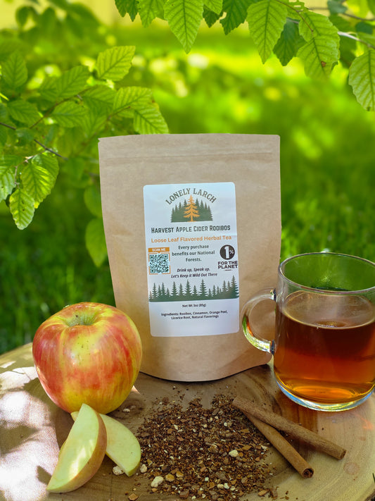 Lonely Larch Harvest Apple Cider Rooibos with apple, cup of tea, and cinnamon sticks on a wooden surface with green leaves in the background.